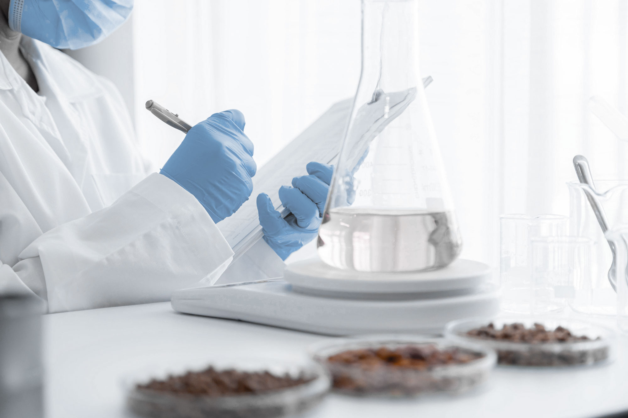 person's hands in lab with gloves writing on clipboard with beaker and product in foreground